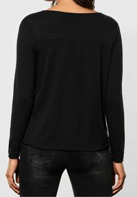 Frau in einem schlichten, schwarzen Langarmoberteil und dunklen Jeans, von hinten vor einem schlichten, hellen Hintergrund gezeigt.