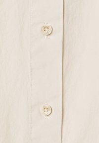 Hellbeiges Hemdmaterial mit zwei runden Knöpfen mit Vierloch-Design, sauber entlang einer geraden Naht angenäht.