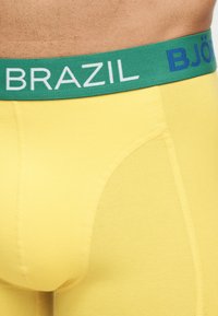 Κοντινό πλάνο κίτρινων εσωρούχων τύπου μποξέρ με πράσινη μέση που φέρει την επιγραφή "BRAZIL", φορεμένα σε ανδρικό σώμα.
