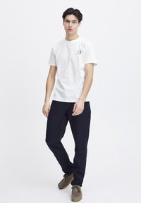 Hvit bomull t-skjorte med korte ermer og en liten svart logo på brystet, kombinert med mørkeblå jeans og beige sko.