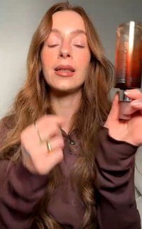 Donna con lunghi capelli castani che tiene e presenta una bottiglia cosmetica di ambra scura, indossa un top marrone a maniche lunghe su uno sfondo uniforme.