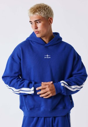 Jeune homme aux cheveux blonds courts portant un sweat à capuche bleu avec des bandes blanches sur les manches, joignant les mains, regardant à gauche.