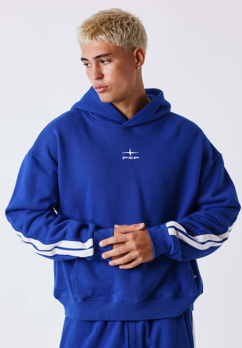 Jeune homme aux cheveux blonds courts portant un sweat à capuche bleu avec des bandes blanches sur les manches, joignant les mains, regardant à gauche.