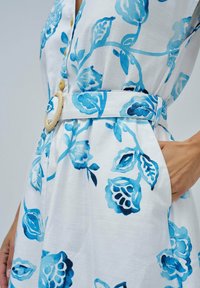 Robe fleurie en tissu blanc avec des motifs de fleurs bleues. Comprend une large ceinture texturée avec une grande boucle ronde.