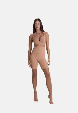 Vrouw staat blootsvoets tegen een witte achtergrond en draagt een huidkleurige beha en hoge shapewear-shorts die tot halverwege de dij reiken.
