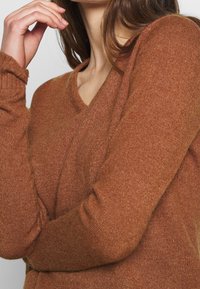 Brun stickad tröja med v-ringad design och ribbade ärmslut, som har en mjuk textur och varm ton. Har lång ärm och en avslappnad passform.