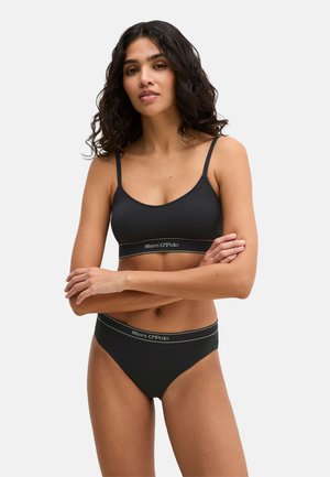 Frau mit lockigem, dunklem Haar, die einen schwarzen, gerippten Bralette und passende schwarze Slips mit Marc O'Polo-Logo auf den Bändern trägt.