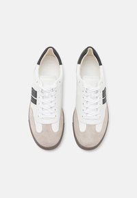 Λευκά δερμάτινα αθλητικά παπούτσια με γκρι σουέτ λεπτομέρειες, μαύρο τακούνι, λευκές επίπεδες κορδόνες και μαύρη λωρίδα κατά μήκος των πλευρών. Ελαστική σόλα.