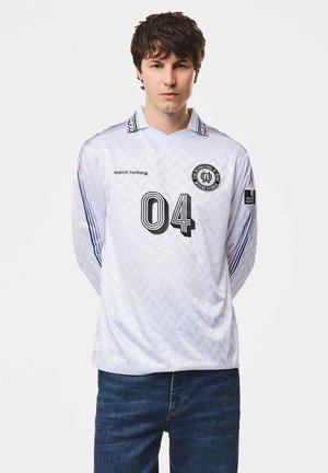 Joven con una camiseta deportiva blanca de manga larga con el número 04, texto "admit nothing," y vaqueros azul oscuro sobre un fondo liso.