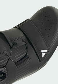 Zapato de ciclismo negro con un diseño elegante, que presenta perforaciones para la transpirabilidad, dos cierres de velcro y detalles texturizados en la parte superior.