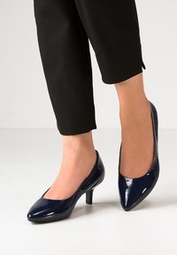 Escarpins en cuir verni marine à bout pointu et talon fin. Portés avec un pantalon noir cropped, mettant en valeur une apparence élégante et soignée.