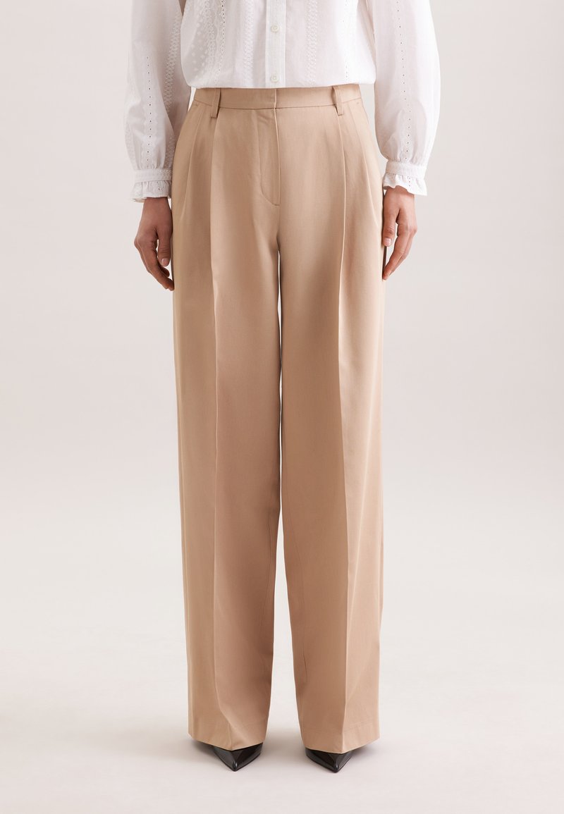 Femme portant un pantalon ample beige et un chemisier blanc à manches longues, debout avec les mains détendues sur les côtés.