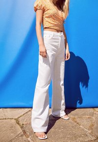 Jeans de pierna ancha blanca, combinados con una blusa corta de flores naranjas. Tejido suave, corte entallado, con un fondo azul sólido.