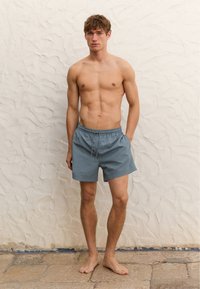 Pantaloni da bagno grigi con cintura elastica e dettaglio con cordoncino, caratterizzati da una vestibilità rilassata. Il modello è a piedi nudi contro un muro testurizzato.