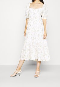 Vestido floral blanco con mangas abullonadas, escote cuadrado, cintura fruncida y falda en capas. Llevado con sandalias blancas de tiras.