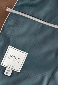 Slim fit sako s podšívkou v odtieni teal s geometrickým vzorom, s bielou etiketou s čiernym textom uvádzajúcim veľkosť a značku. Viditeľný hnedý vonkajší materiál.
