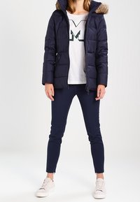 Donna che indossa un giubbotto imbottito blu navy con cappuccio rifinito in pelliccia, una t-shirt bianca con stampa, pantaloni slim blu navy e sneakers bianche, in piedi su uno sfondo bianco.