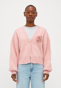 Rozā kardigāns ar brīvu piegriezumu, V veida kakla izgriezumu, ribotām malām un auduma ziedu akcentu uz krūts. Valkāts virs pelēkas krekla un zilu džinsu.