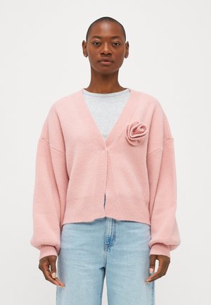 Pink cardigan with a loose fit, v-neck, ribbed edges, and a fabric flower accent on the chest. Worn over a gray shirt and blue jeans.