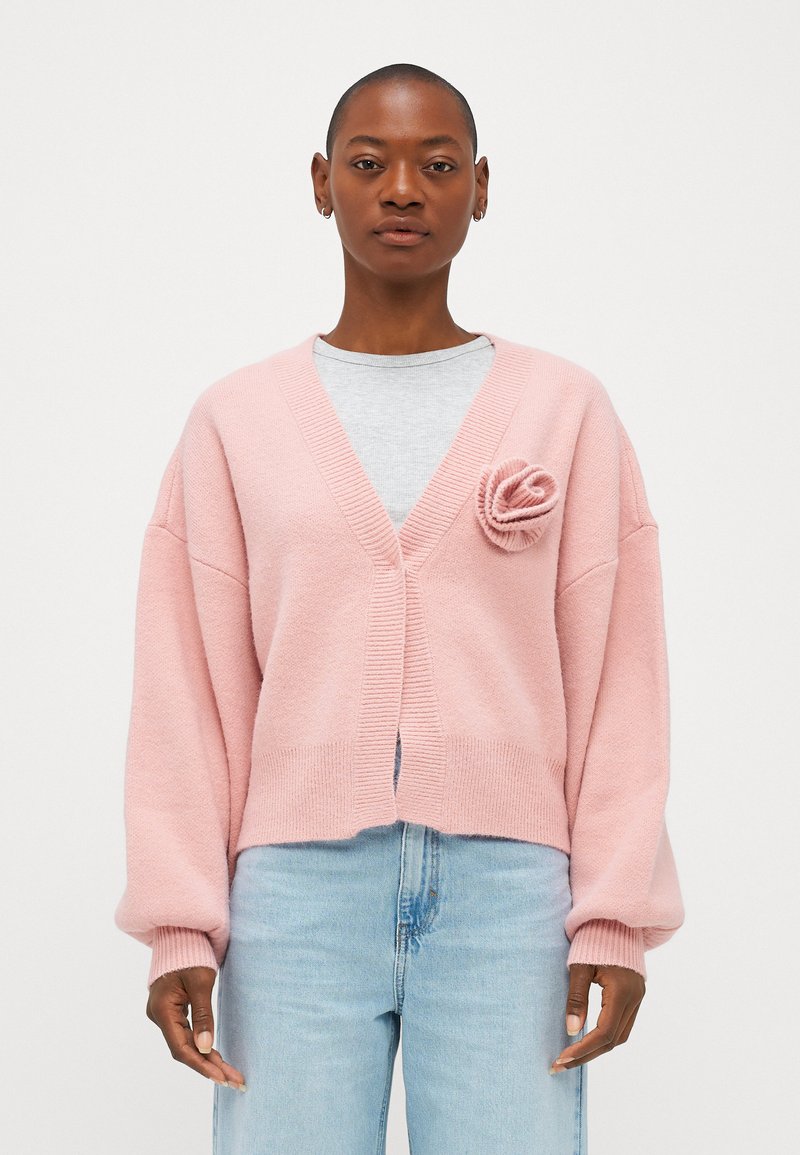 Rozā kardigāns ar brīvu piegriezumu, V veida kakla izgriezumu, ribotām malām un auduma ziedu akcentu uz krūts. Valkāts virs pelēkas krekla un zilu džinsu.