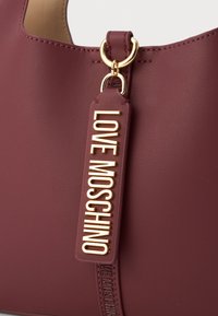 Burgundia faux nahast käekott, millel on kuldne logo silt, ümar kuju, sile tekstuur ja kuldsed detailid.