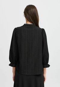 Blouse noire à rayures verticales, coupe ample, manches bouffantes et poignets à volants. Finition avec un col orné de dentelle pour plus de détail.