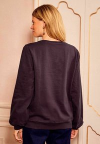 Temno siva majica s krojem okroglega vratu, dolgimi rokavi in rebrastimi manšetami, v kombinaciji z modrimi hlačami. Gladka tekstura tkanine z minimalnimi šivi.