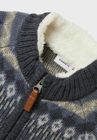 Gruby sweter dzianinowy z niebiesko-szaro-beżowym wzorem, żebrowanym kołnierzem, brązowym skórzanym uchwytem zamka błyskawicznego oraz białym futerkiem na podszewce kołnierza.