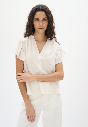 Beige blouse met korte mouwen en een V-hals, met een gestructureerde stof en geplooide accenten bij de schouders. Draag het met lichtgekleurde broeken.