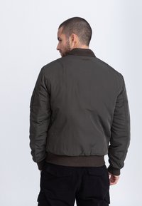 Oliwkowa bomberka wykonana z gładkiej tkaniny, z prążkowanymi mankietami i krawędzią oraz małym haftowanym detalem na plecach.