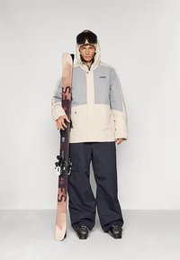 Tânărul îmbrăcat într-o jachetă de schi gri și bej și pantaloni negri ține schiurile în poziție verticală, purtând ochelari de schi pe cap și bocanci de schi.