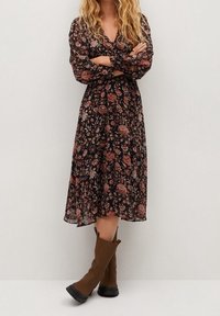Blumenkleid in Midi-Länge mit schwarzem Grund, das mit pinken und orangefarbenen Blumen verziert ist. Lange Ärmel, ein V-Ausschnitt und kombiniert mit braunen kniehohen Stiefeln.