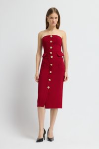 Rotes trägerloses Kleid mit tailliertem Oberteil, goldenen Knopfakzenten, zwei vorderen Taschen und einem Schlitz am Knie. Das Modell trägt schwarze High Heels.