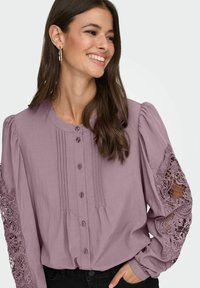 Naeratav naine, kandes lavendlivärvi nööbitavat pluusi pitsiliste lõigetega varrukatel ja musti pükse, käsi taskus, ühevärvilise taustaga.