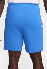 Man som bär ljusblå knälånga shorts, stående, synlig från midjan till mitt lår mot en enfärgad bakgrund.