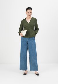 Žena u maslinasto zelenoj bluzici s preklopom, širokim plavim trapericama i bež slingback cipelama s crnim prstima, drži malu bež torbicu.