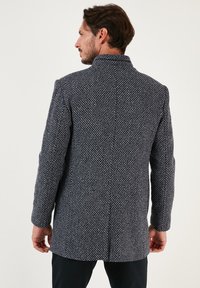 Cappotto blu navy in una lavorazione a maglia lavorata con un colletto alto e maniche lunghe. Presenta una linea dritta e un dettaglio di spacco posteriore.