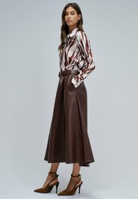 Falda midi de cuero marrón con un patrón de cocodrilo texturizado, combinada con una blusa blanca con un estampado abstracto marrón y tacones marrones.