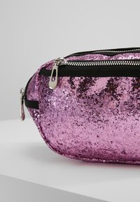 Růžová ledvinka zdobená flitry s oblým tvarem, vybavená černým zipem a textilním lemem. Třpytivý povrch odráží světlo a zdůrazňuje texturovaný design.