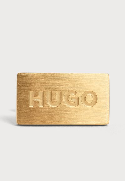 Четка златна правоъгълна метална плоча с думата "HUGO" вдълбана в центъра върху светъл фон.