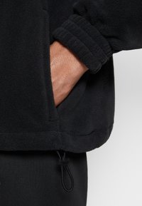 Schwarzer Fleece-Pullover mit gerippten Bündchen und Seitentasche. Elastischer Kordelzug am Saum. Weiche Textur mit matter Oberfläche.