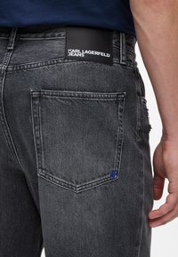 Crne traperice sivo prane boje, s kožnom oznakom "Karl Lagerfeld Jeans" i plavim šivom na stražnjem džepu.