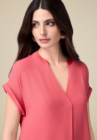 Blusa rosa corallo con scollatura a V e maniche corte arrotolate. Tessuto leggero con una consistenza liscia, caratterizzato da una leggera lucentezza e linee pulite.