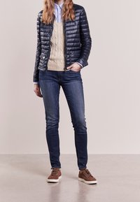 Nainen, jolla on päällään tummansininen topattu takki vaaleanbeige villapaita, siniset farkut ja ruskeat mokkanahkatennarit, seisoo yksi käsi taskussa.