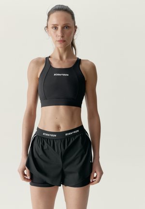 Mujer en forma con el cabello recogido, lleva un sujetador deportivo Born to Run negro y shorts, de pie frente a un fondo claro y liso.