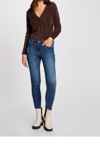 Bruine, pluizige trui met een V-hals en drie knopen, gecombineerd met blauwe skinny jeans met een vervaagd patroon en lichte slijtage. Witte enkellaarsjes.