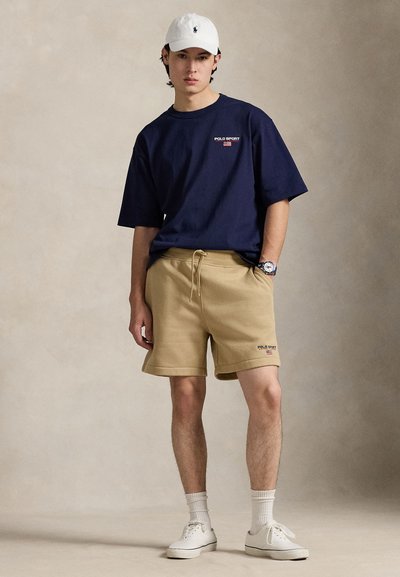 Μπλε βαμβακερό μπλουζάκι με κοντά μανίκια και λογότυπο «Polo Sport». Μπεζ βαμβακερά σορτς με κορδόνι. Λευκό καπέλο μπέιζμπολ και αθλητικά παπούτσια.