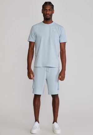 Mężczyzna z warkoczami, ubrany w jasnoniebieską koszulę z krótkim rękawem i pasujące krótkie spodnie typu cargo, stojący na jednolitym jasnym tle.