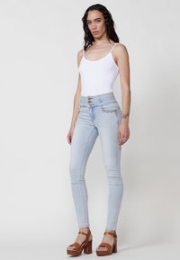 Vaqueros ajustados de tiro alto en un azul claro, con acentos de botones dorados y costuras decorativas. Combinados con una camiseta sin mangas blanca ajustada y sandalias de tacón marrones.