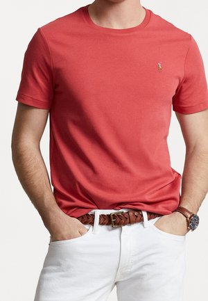 Mężczyzna noszący dopasowany czerwony t-shirt z krótkim rękawem z małym logo polo, białe spodnie, brązowy pleciony skórzany pasek i brązowy zegarek na rękę.